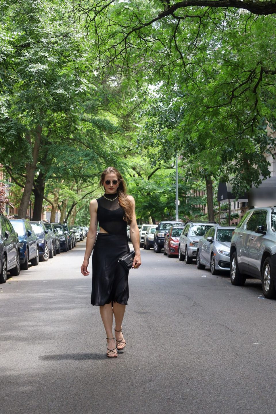Walking toward the camera down a tree lined street in a sheer black top and black slip skirt.