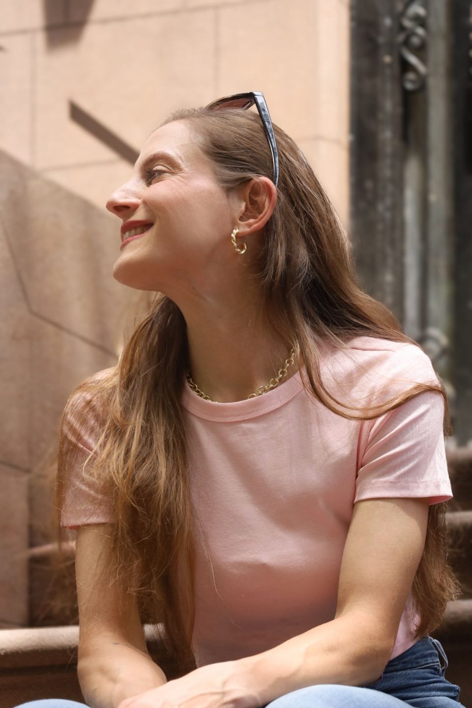 Seated and laughing in profile, pink tee, sunglasses pushed up into her hair.