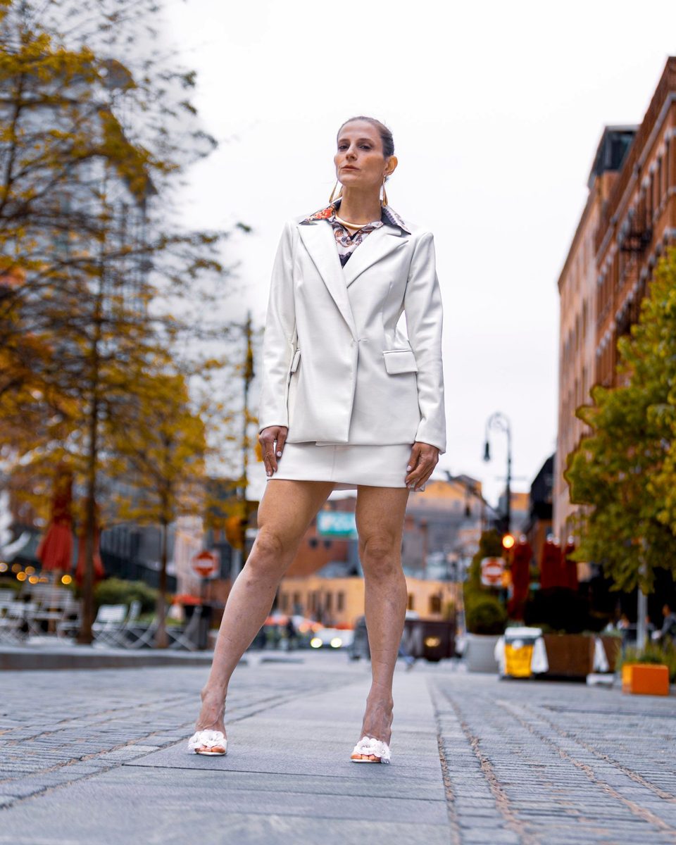 Low angle, standing on cobblestones in the ivory suit, autumn trees behind her.
