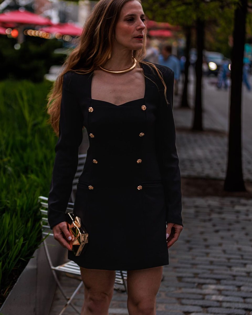 A black double breasted mini dress with gold buttons, gold collar necklace, on a Manhattan street.