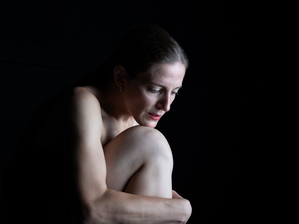Studio portrait on black, knees drawn up, looking down.