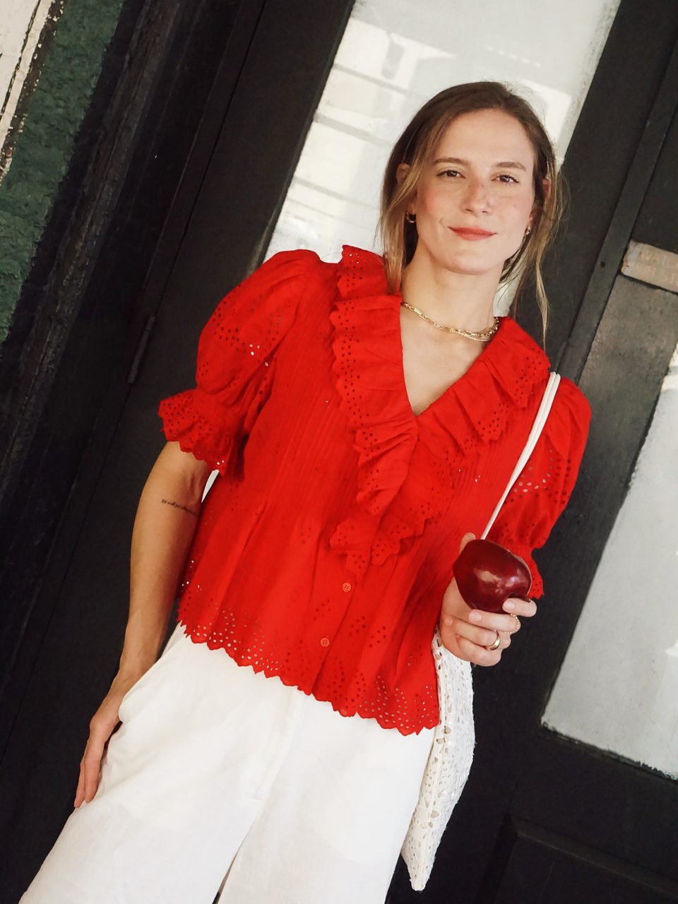 A red ruffled eyelet blouse and white trousers, holding a red apple, leaning in a doorway.
